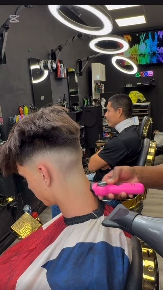 Un barbero le hace un corte de pelo degradado a un joven en una barbería, usando una máquina cortadora y un secador de pelo.
