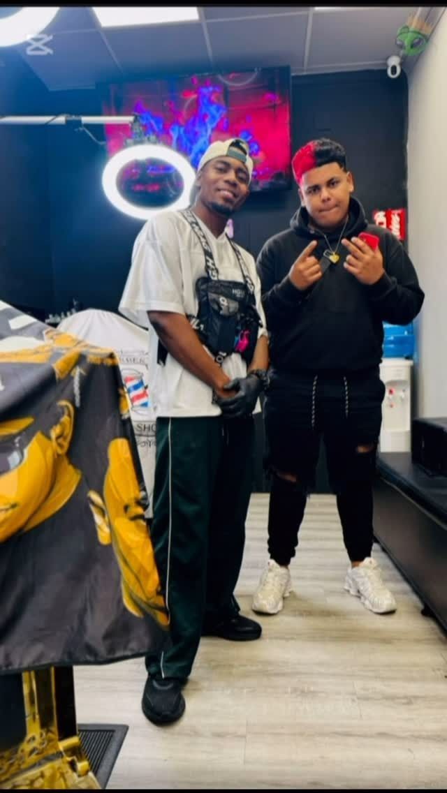 Dos hombres posan en una barbería. Uno lleva una camisa blanca y el otro una sudadera negra con capucha, ambos con un look informal.