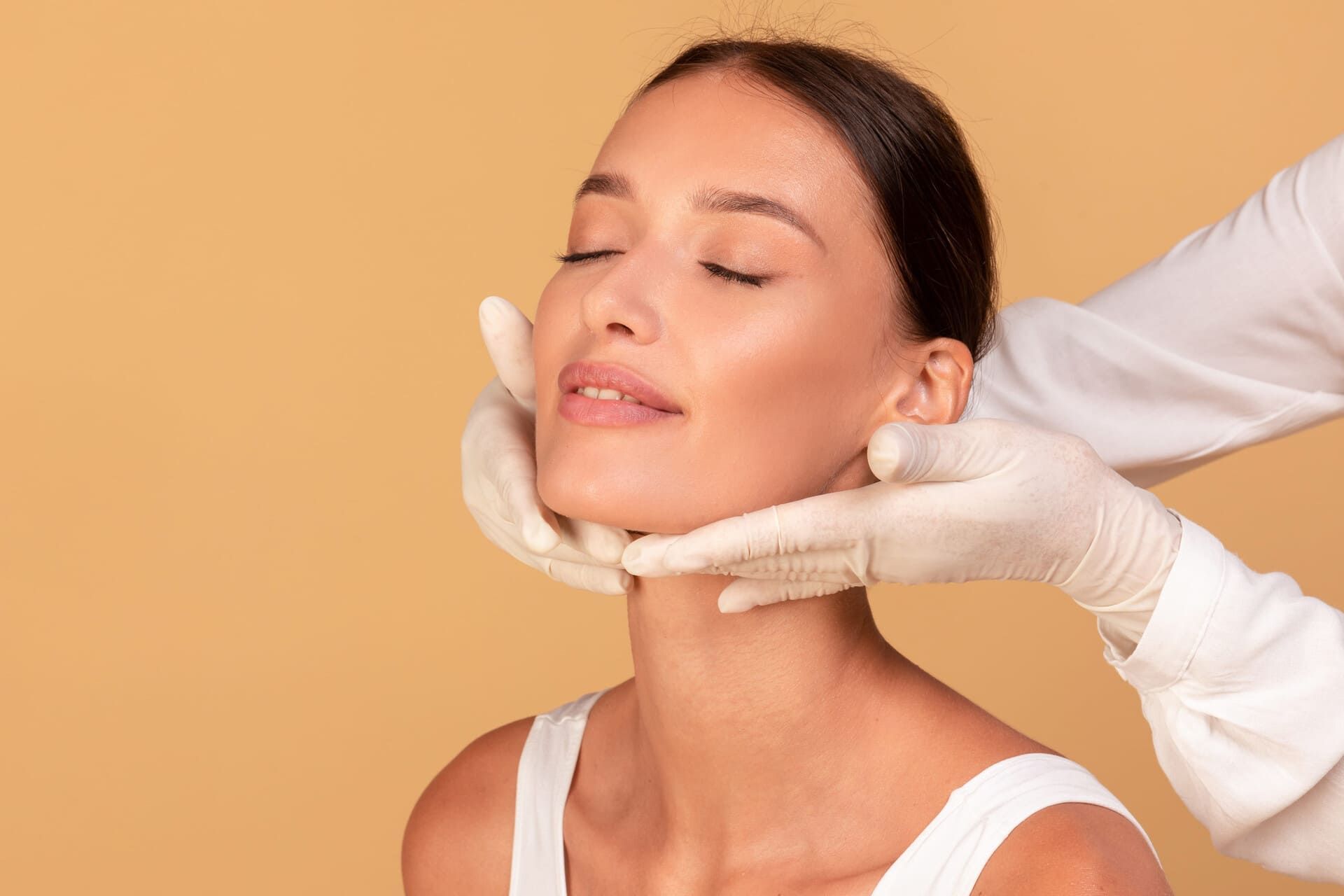 Tratamiento facial cosmético en una mujer con manos enguantadas que levanta suavemente su barbilla sobre un fondo color melocotón.