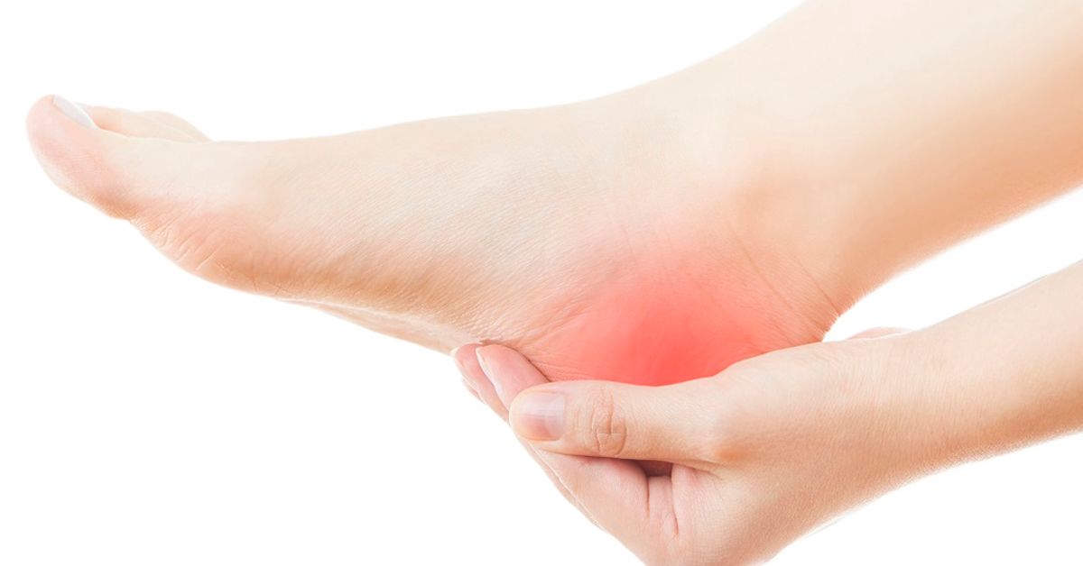 Mano sosteniendo el pie, resaltado rojo en el talón, lo que indica dolor.