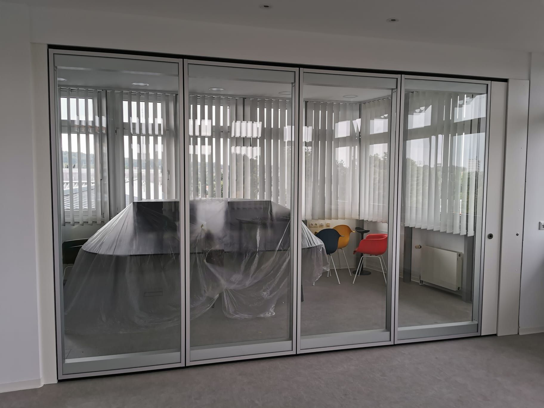 Durch gläserne Falttüren gelangt man in einen Raum mit einem abgedeckten Tisch, Stühlen und Jalousien.