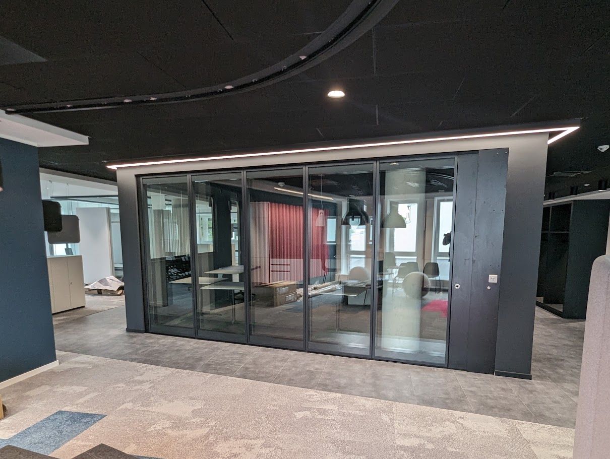 Büroraum mit Glaswänden und dunklem Rahmen. Inklusive geschlossener Schiebetür, schwarzer Decke und einigen sichtbaren Büromöbeln.
