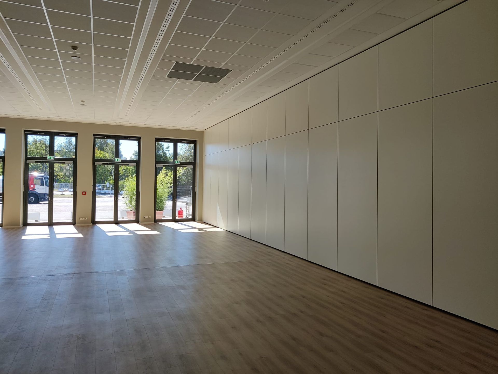 Leerer, sonnendurchfluteter Raum mit Holzboden, Fenstern und hellbeiger Holzvertäfelung an einer Wand.