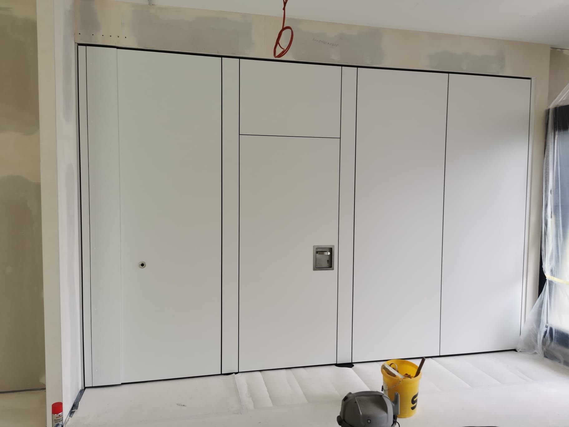 Eine Wand ist mit weißen Paneelen verkleidet und verfügt über eine kleine Tür mit Griff und Schloss. Auf dem Boden stehen ein Eimer und Werkzeug.