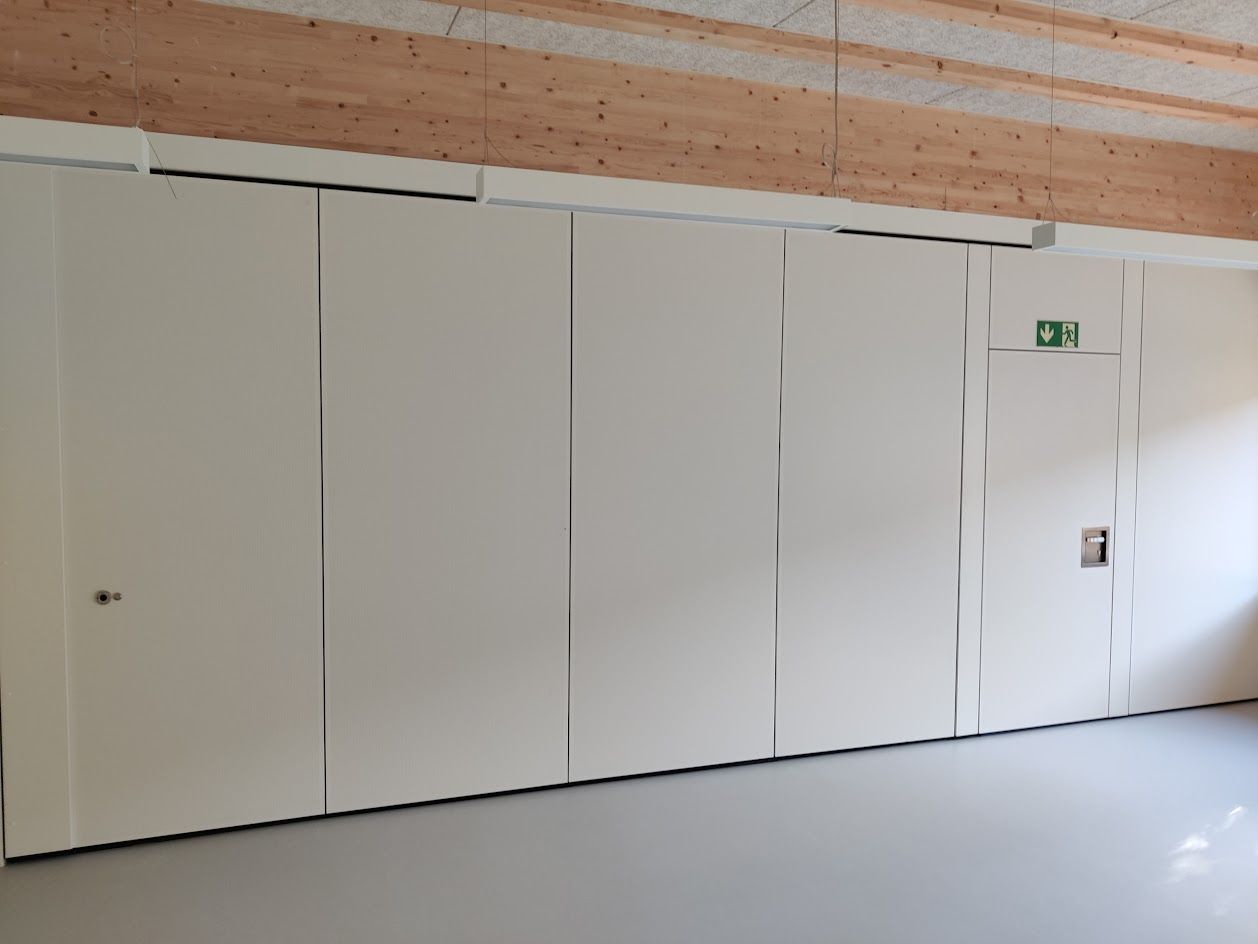 Weiße Faltwand mit Tür in einem Raum mit hellgrauem Fußboden und Holzdecke.