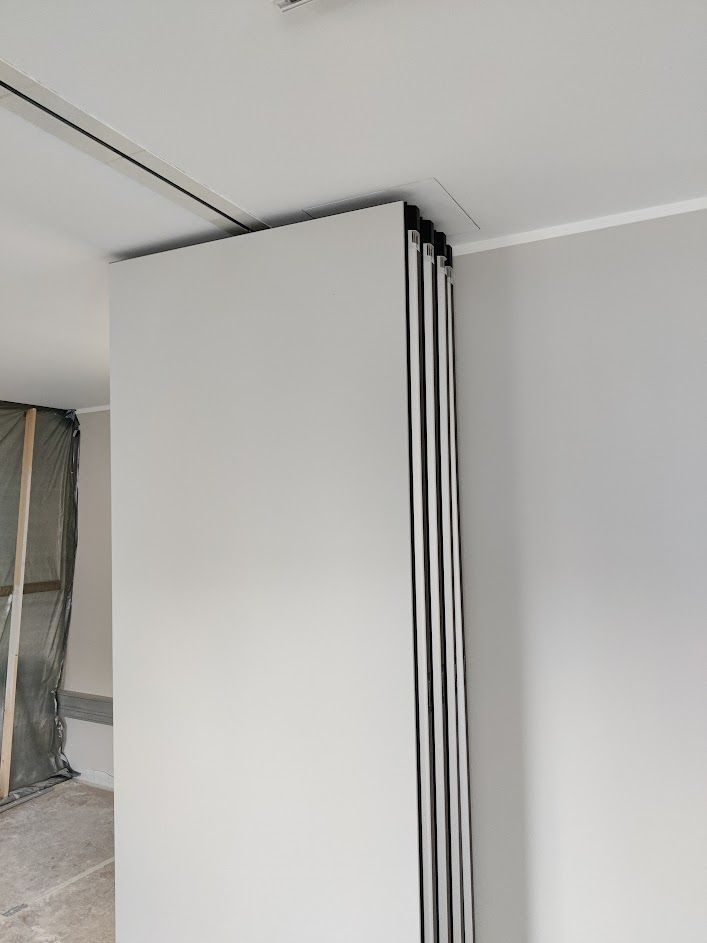 Faltbare Trennwand, teilweise geschlossen, weiße Paneele, schwarze Kanten, in einer Ecke installiert, Deckenschiene sichtbar.