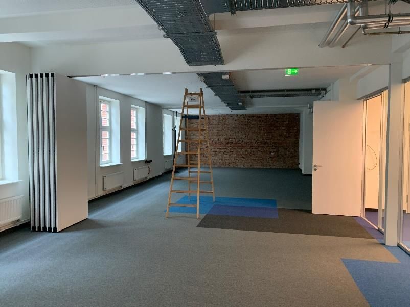 Leerer Raum mit Fenstern, freiliegender Ziegelwand, einer Leiter und Kabelrinnen an der Decke.