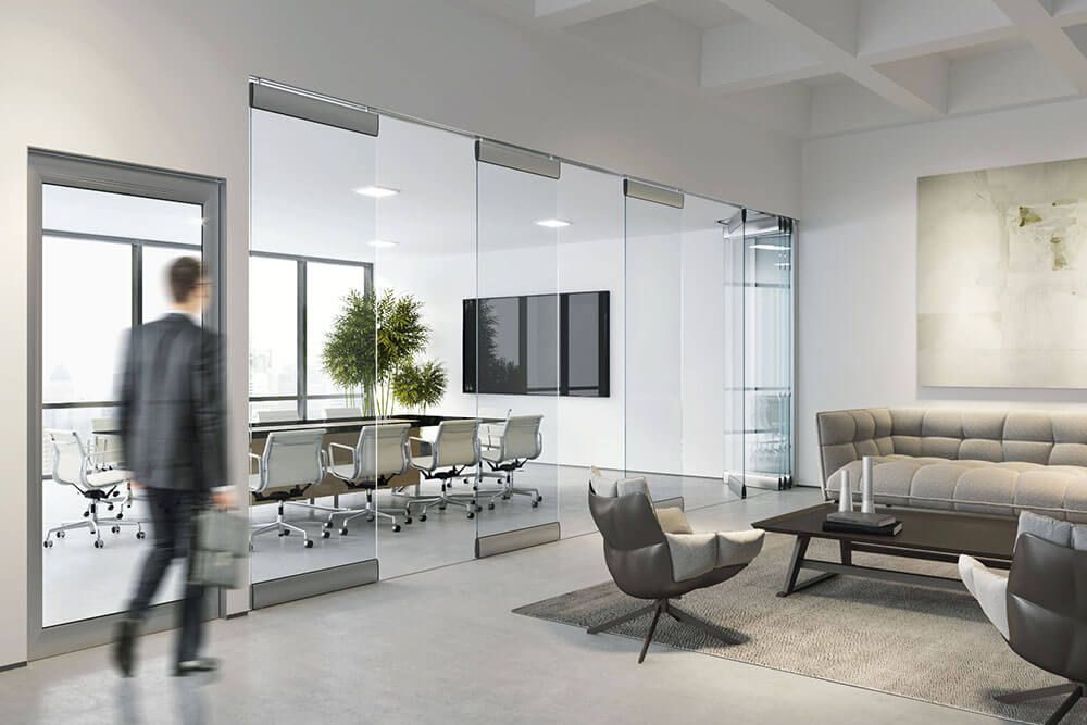 Ein Mann im Anzug geht an einem Büro mit Glaswänden vorbei, in dem ein Konferenztisch, Stühle und eine Sitzecke stehen.