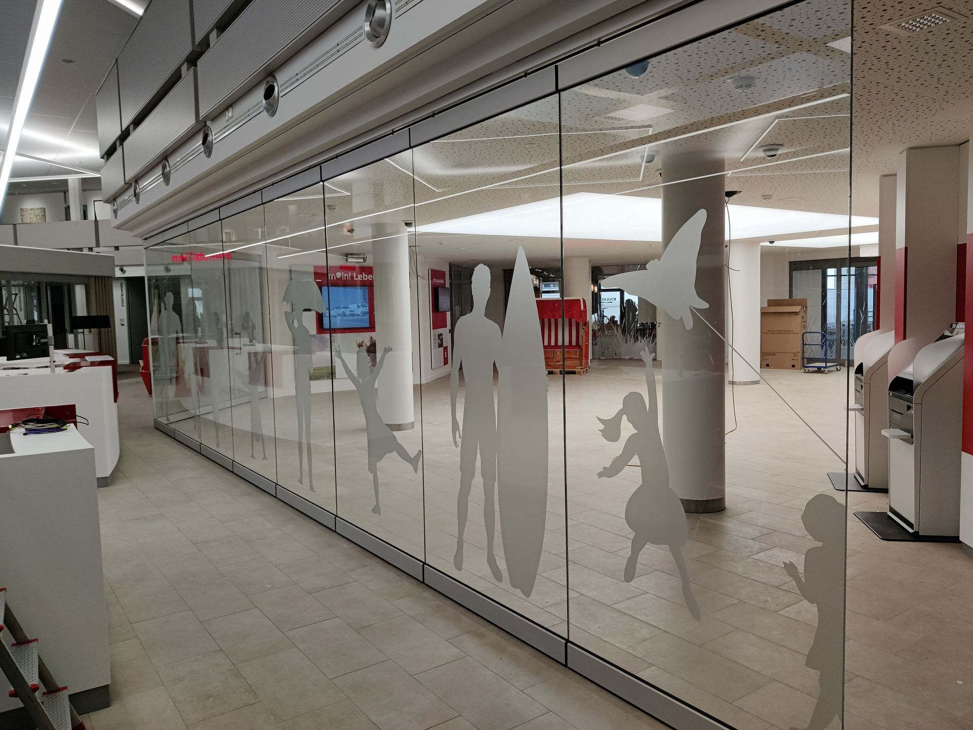 Glastrennwand mit mattierten Silhouettenfiguren in einem modernen Interieur, möglicherweise einer Bibliothek oder einem öffentlichen Raum.