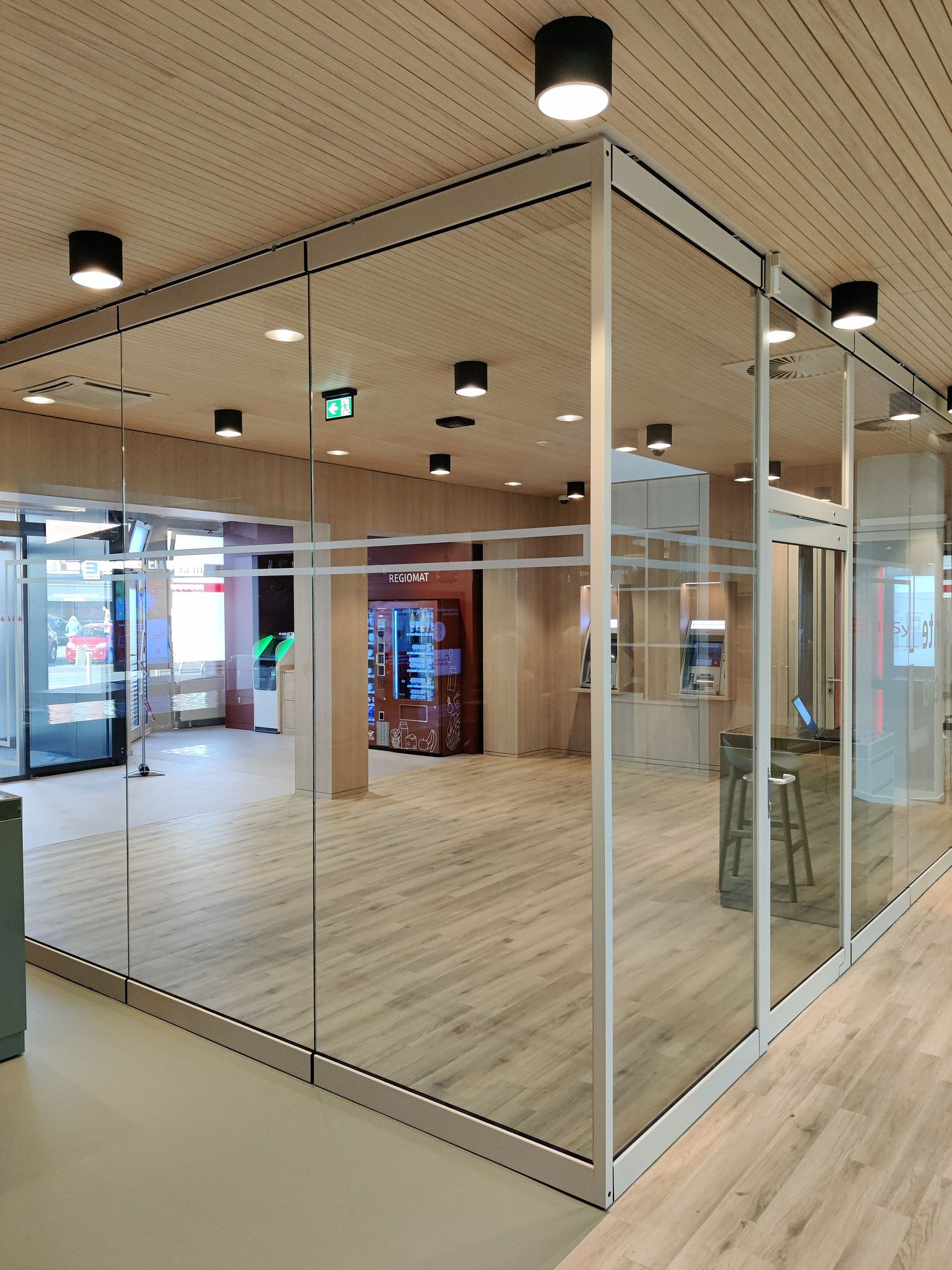 Ein verglaster Raum mit Holzboden und -decke sowie mehreren dunklen Deckenleuchten. Im Inneren befinden sich ein Kiosk und einige Sitzgelegenheiten.