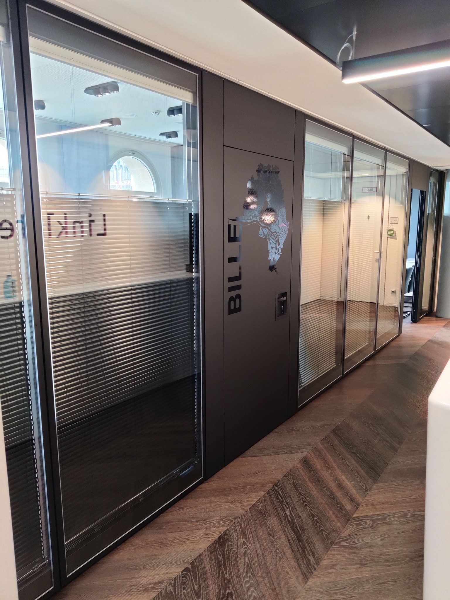 Flur mit verglasten Büros. Dunkelgraue Türen und Rahmen. Brauner Teppichboden im Fischgrätmuster.