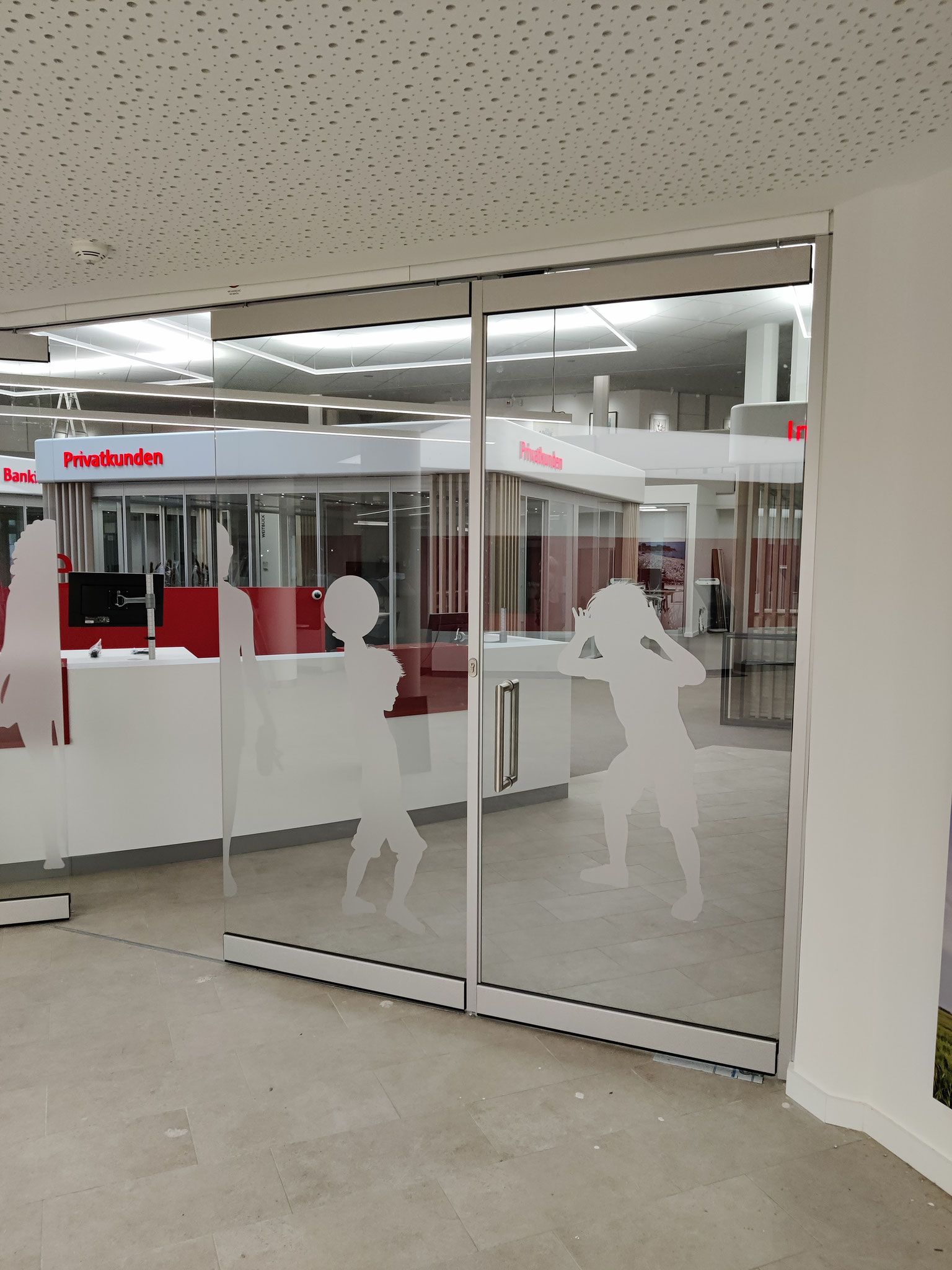 Schiebetür aus Glas mit mattierten Silhouettenfiguren. Führt in einen hellen Innenraum mit roten Akzenten.