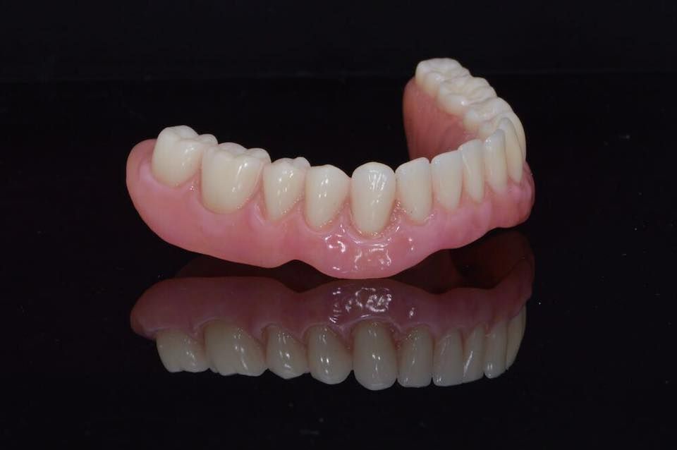 Dentadura superior con base rosa y dientes blancos, reflejados sobre una superficie oscura.