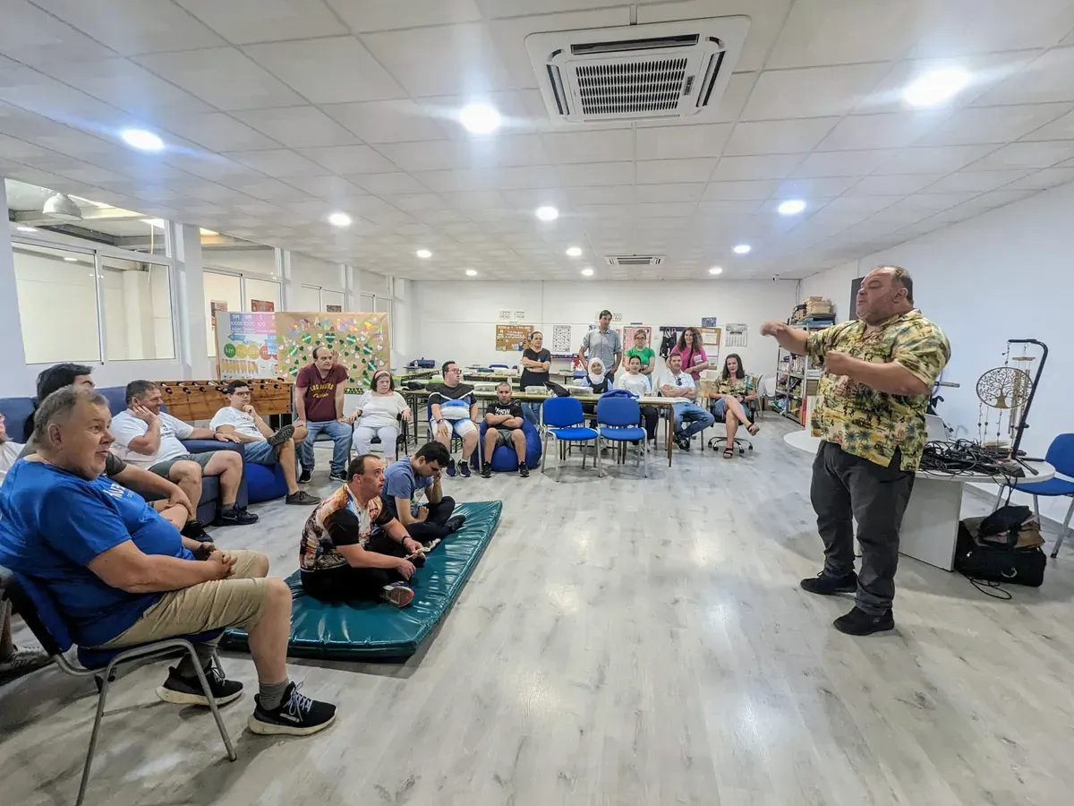 Reunión comunitaria en un salón luminoso, con los asistentes sentados observando a un orador de pie al frente.
