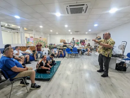 Reunión comunitaria en un salón luminoso, con los asistentes sentados observando a un orador de pie al frente.