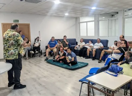 Un grupo de personas está sentado en sofás y sobre una esterilla en una habitación, escuchando a un hombre que está de pie y les habla.