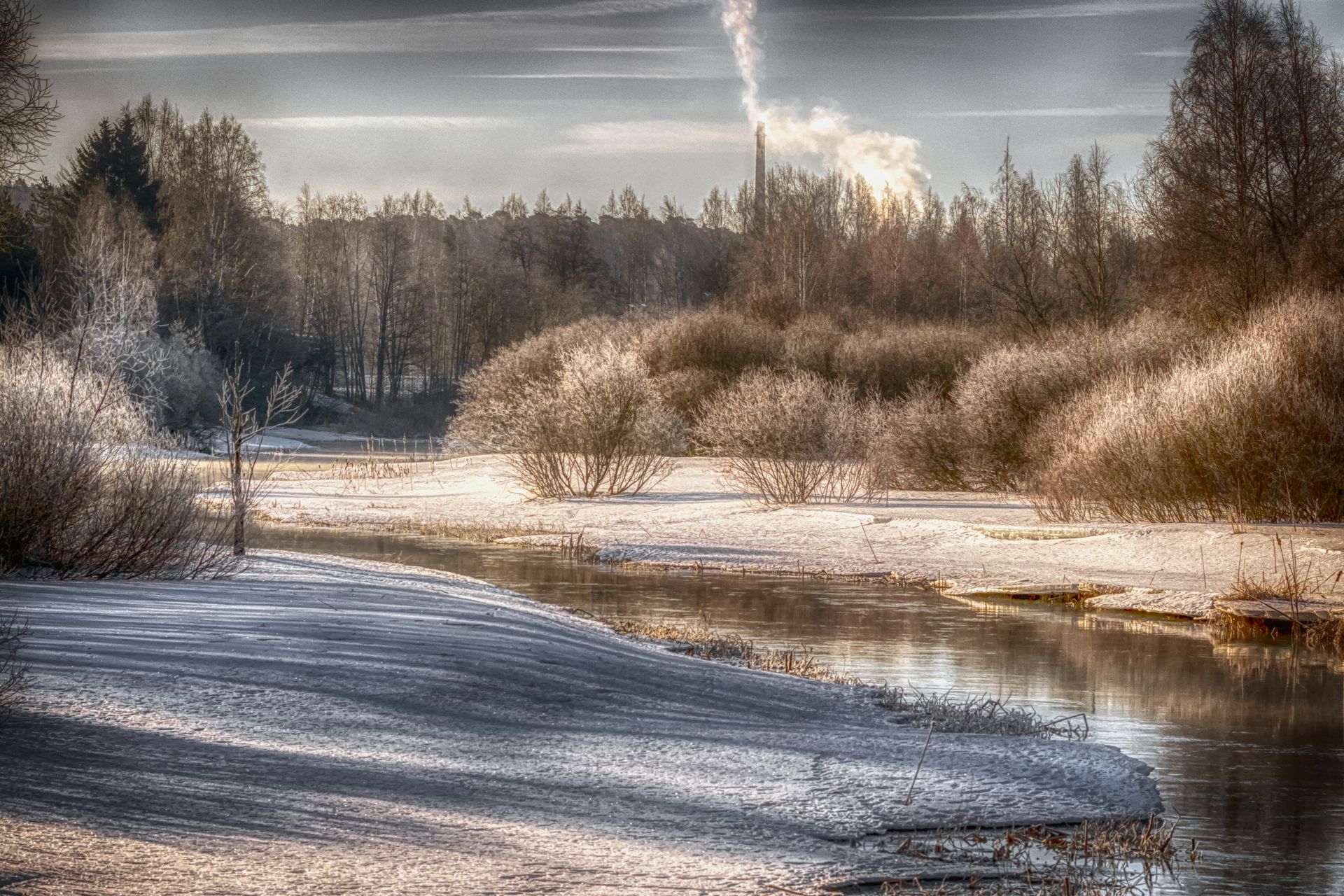 Luminen joenranta auringonnousussa, kuuraiset puut ja kultainen valo heijastuvat tyynestä vedestä.