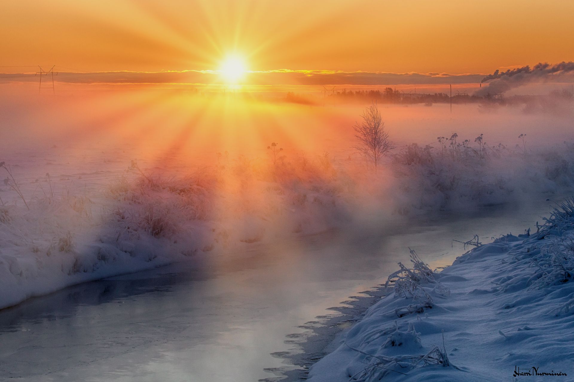 Auringonnousu sumuisen, jäätyneen joen yllä, oranssia valoa ja lumiset rannat