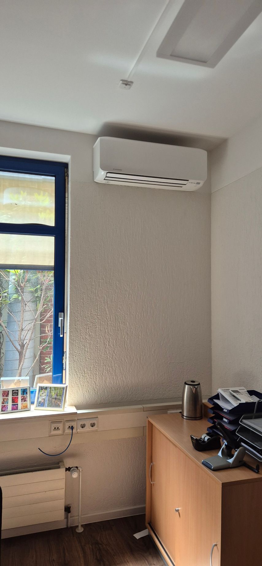 Büroecke mit Fenster, Heizkörper, Wandklimaanlage und einem Holzschrank mit Drucker.