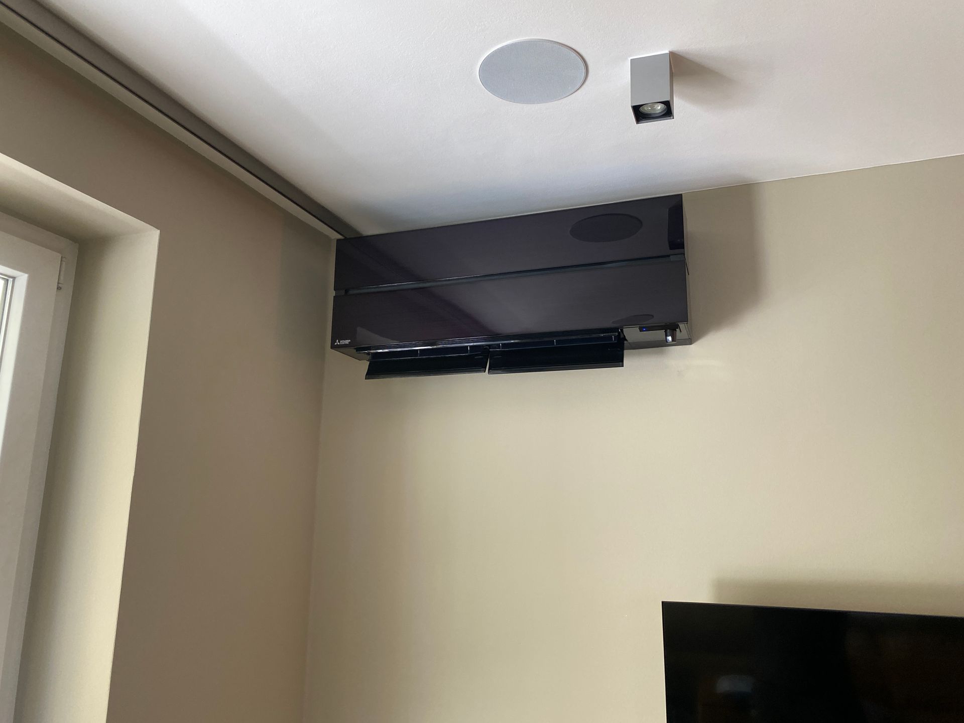 An der Wand montierter Fernseher über einem Eckfenster in einem beigen Zimmer mit Deckenlautsprecher und kleinem Gerät