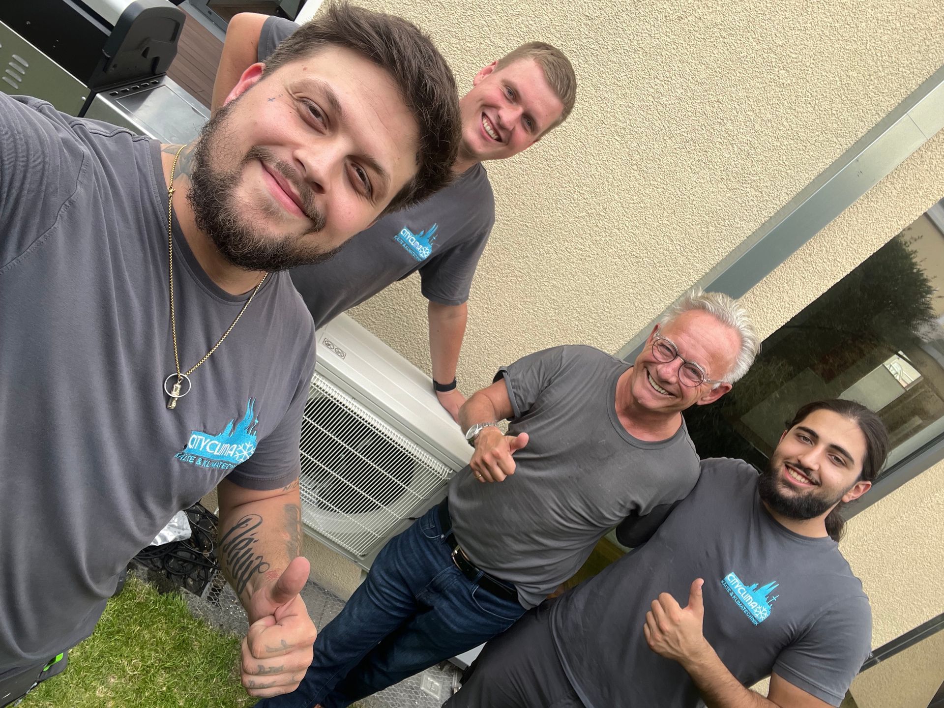 Vier Personen in passenden grauen Hemden lächeln im Freien und machen ein Selfie in der Nähe eines Gebäudes.