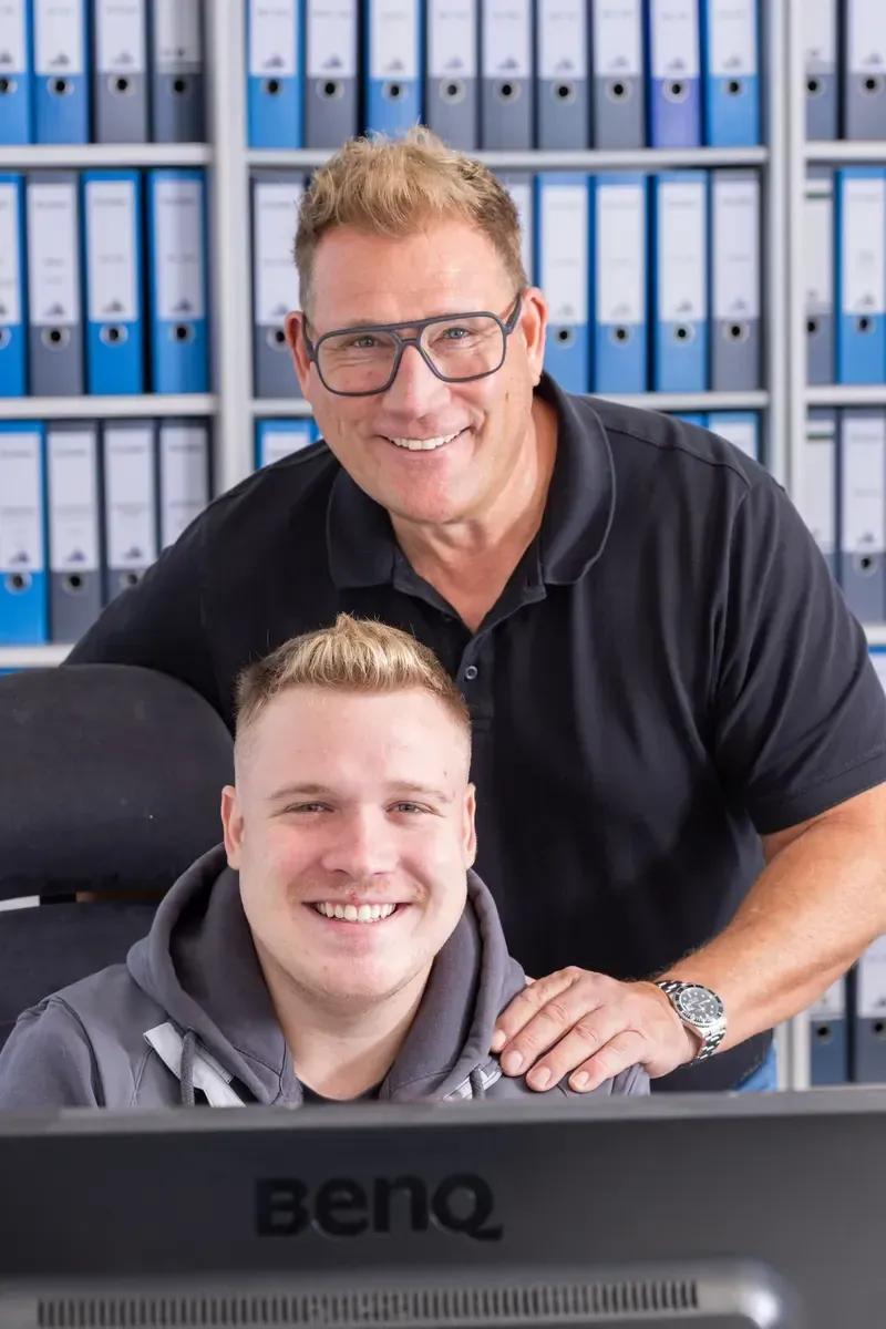 Zwei lächelnde Kollegen an einem Schreibtisch in einem Büro, hinter ihnen in Regalen blaue Ordner.