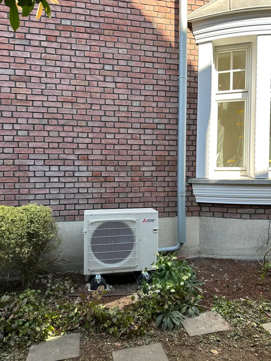 Außenklimaanlage neben einer gemauerten Hauswand und einem Fenster, in der Nähe von niedrigen Sträuchern und Gartensteinen