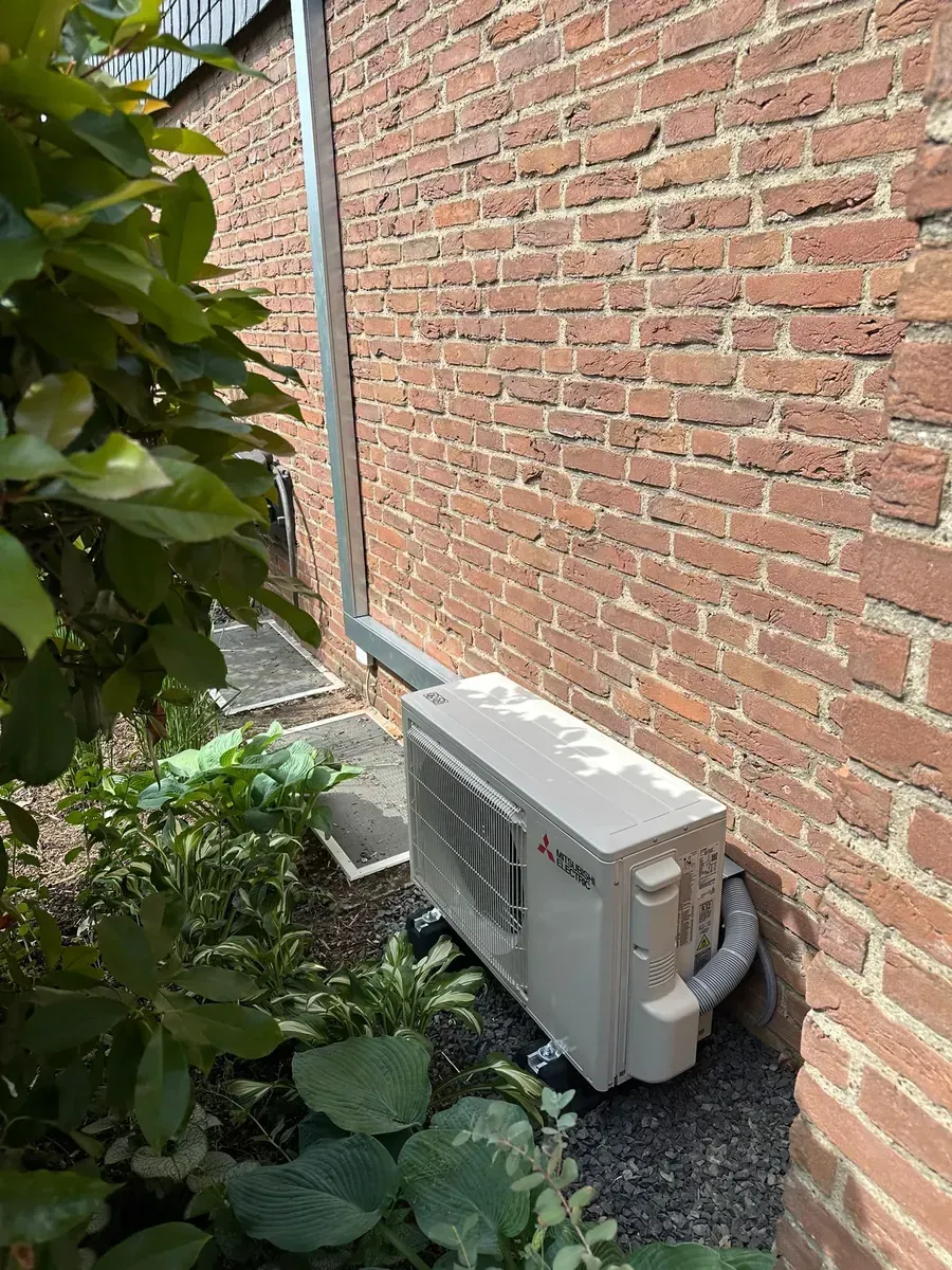 Außenklimaanlage neben einer Backsteinmauer, teilweise umgeben von dichtem Grün.