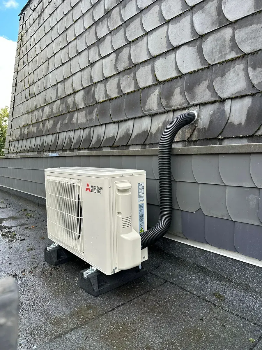 An der Wand montierte Außenklimaanlage neben einer mit Schindeln gedeckten Hauswand auf einem feuchten Dach.