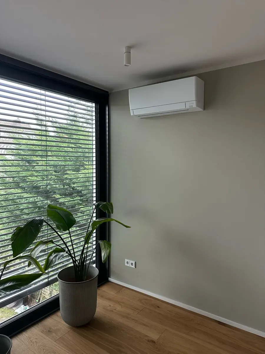 Moderne Zimmerecke mit großen Jalousien, Topfpflanze, Holzboden und wandmontierter Klimaanlage.