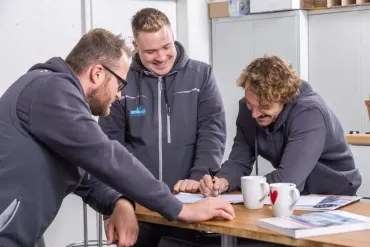 Drei Kollegen in grauen Jacken sitzen in einem Bürotisch und arbeiten gemeinsam an Unterlagen.