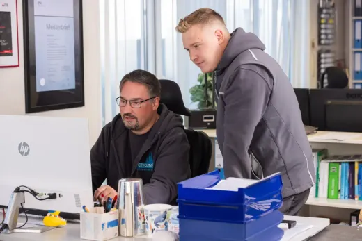 Zwei Kollegen unterhalten sich an einem Schreibtisch mit Computer und blauen Tabletts in einem hellen Büro.