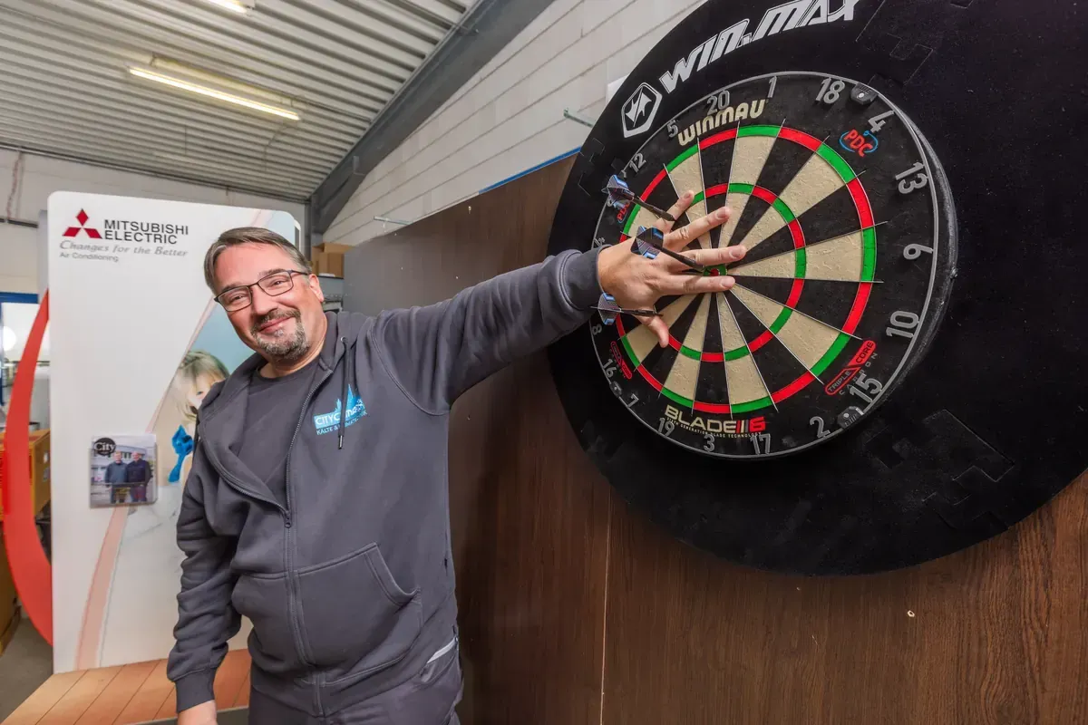 Ein Mann lächelt neben einer Dartscheibe und zeigt in einer Indoor-Arena auf die Mitte.