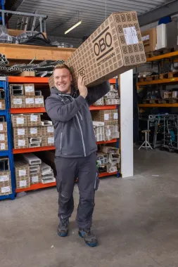 Eine Person hält in einem Lagerhaus einen großen Karton über dem Kopf.