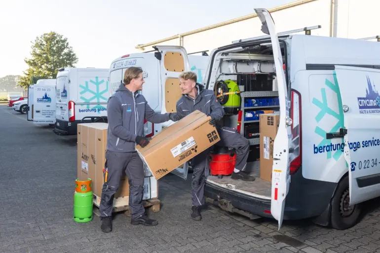 Zwei Arbeiter entladen Kartons aus einem weißen Lieferwagen an einem Auslieferungsort im Freien.