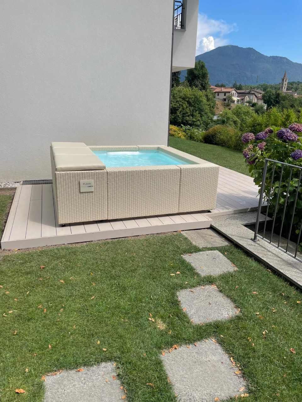 Una vasca idromassaggio beige è situata su una terrazza in legno accanto a un edificio bianco e a un prato verde.