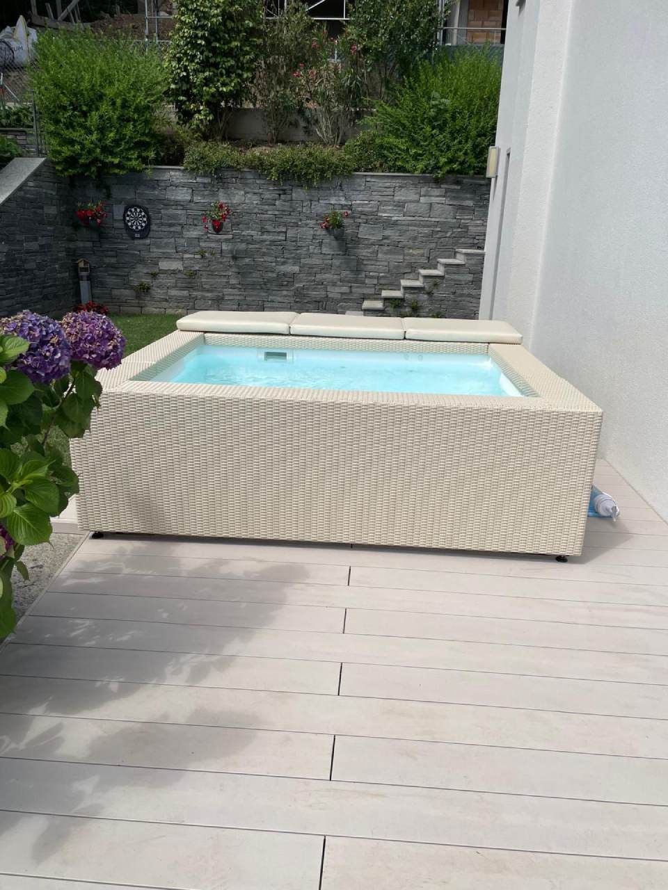 Vasca idromassaggio quadrata su un patio dai colori chiari; sullo sfondo un muro in pietra e delle scale.