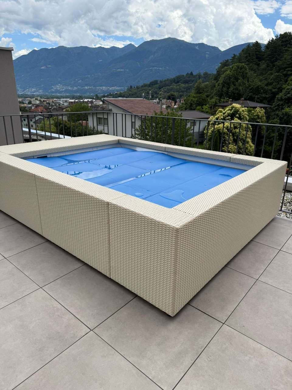 Una vasca idromassaggio rettangolare in vimini bianco con copertura blu, su un tetto piastrellato con vista sulle montagne.