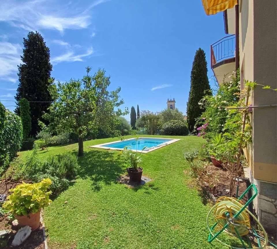 Un cortile con una piscina rettangolare, prato verde, alberi e un edificio con un balcone sotto un cielo azzurro.
