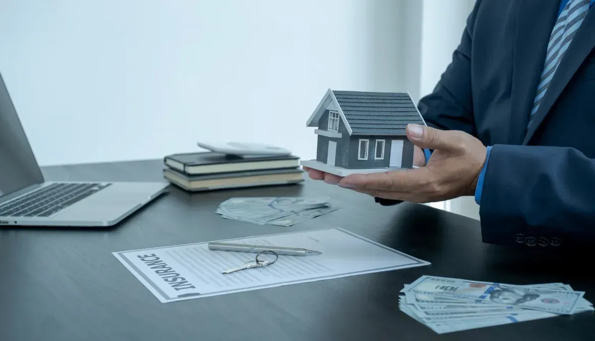 Persona con traje sostiene una maqueta de casa, con papeles, una computadora portátil y dinero sobre un escritorio.