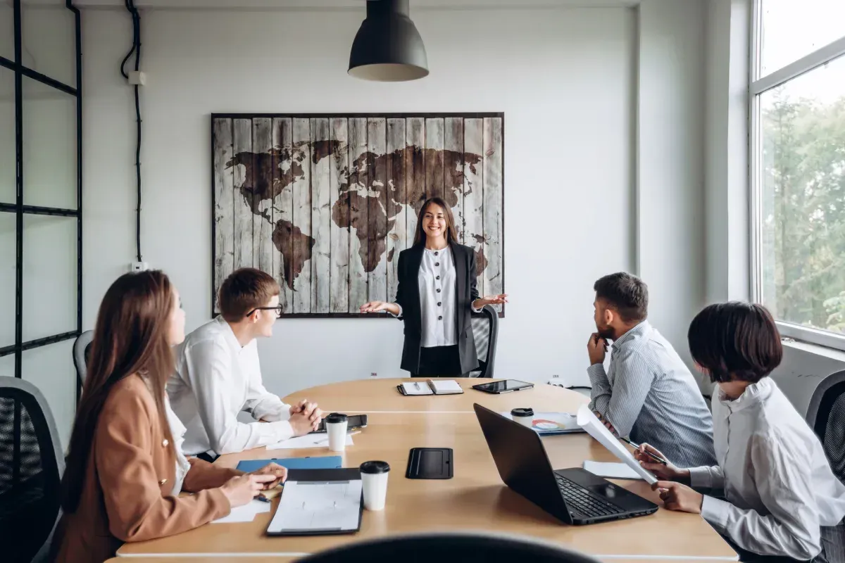 Una mujer presenta en una reunión, de pie junto a un mapamundi de madera. Cuatro personas sentadas alrededor de una mesa