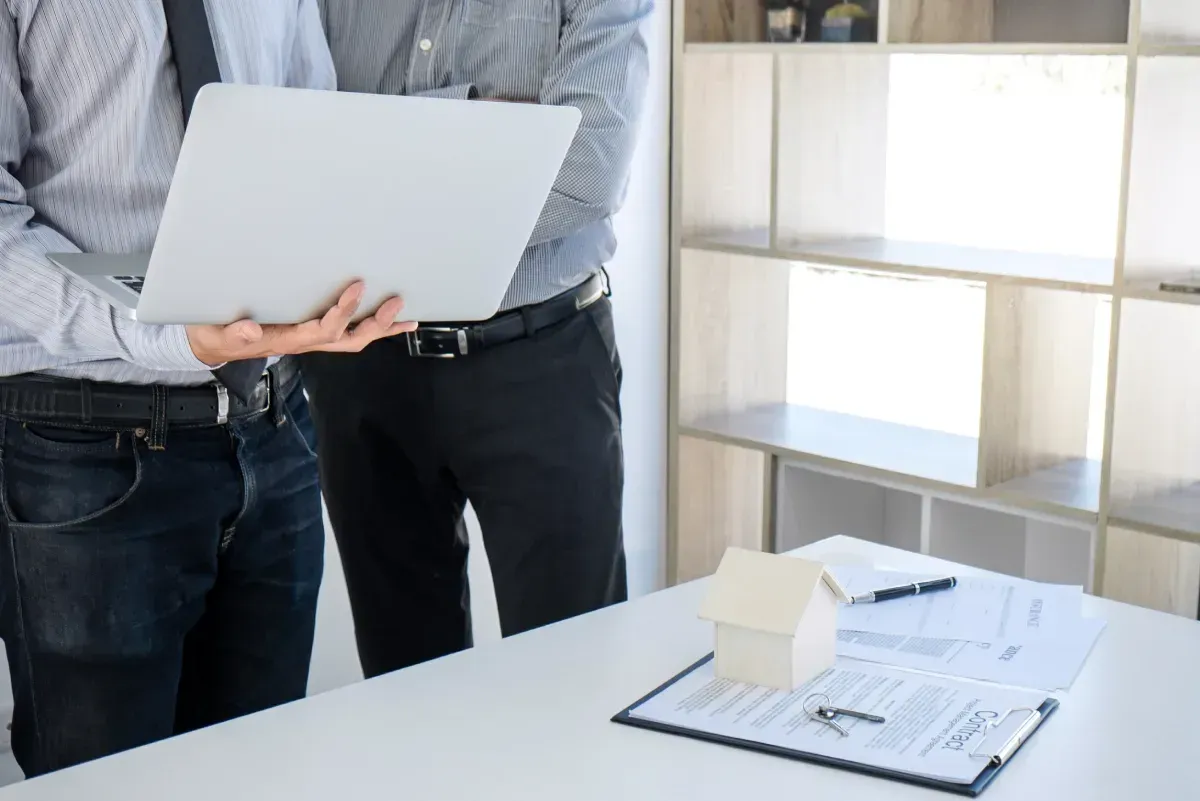 Dos personas revisan una computadora portátil cerca de un escritorio con una maqueta de casa y documentos en una oficina.