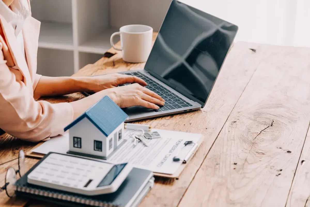 Mujer usando una computadora portátil en un escritorio con un modelo de casa, documentos, calculadora y taza de café.