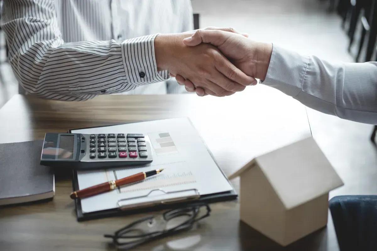 Dos personas se dan la mano mientras comparten papeles y una calculadora, con la posibilidad de cerrar un trato.