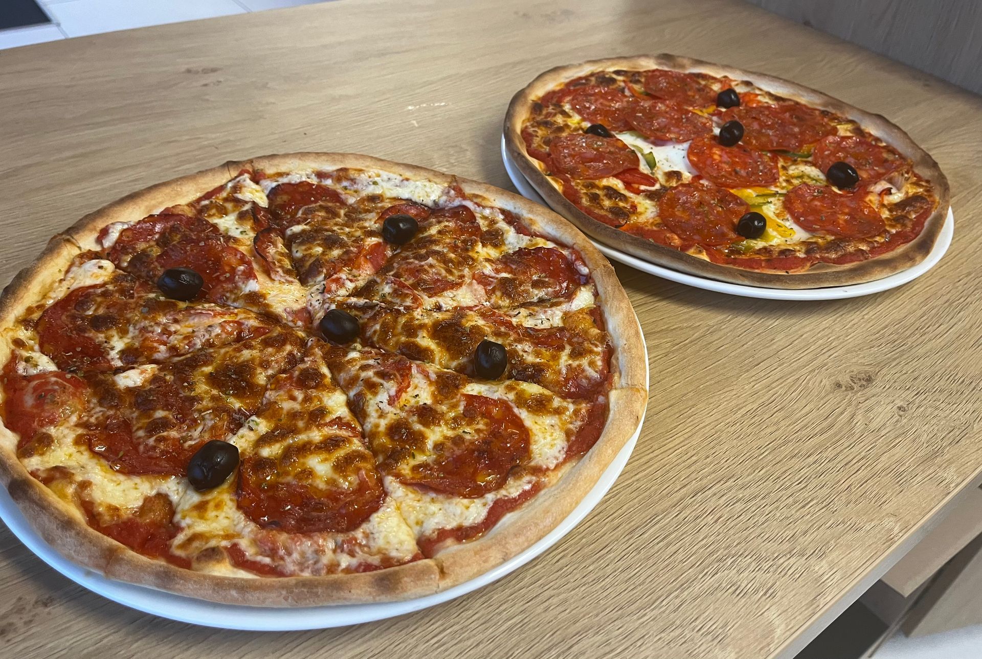 Deux pizzas sur des assiettes blanches posées sur une table en bois, l'une au fromage et aux olives, l'autre au pepperoni et aux olives.
