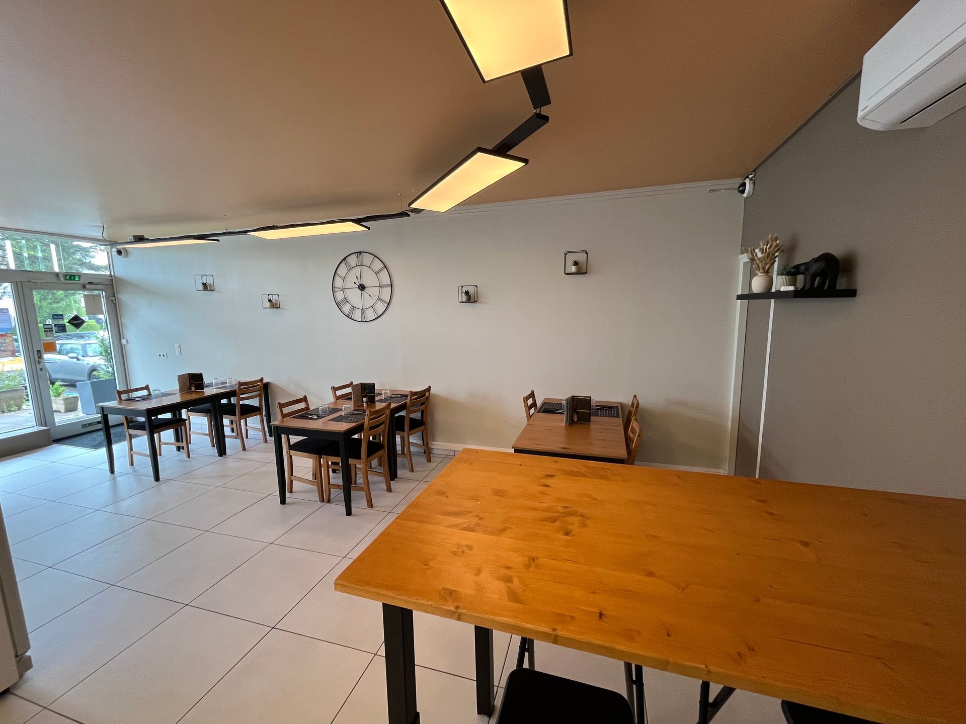 Intérieur d'une pizzeria avec tables en bois, sol carrelé blanc, horloge murale et grande vitrine.