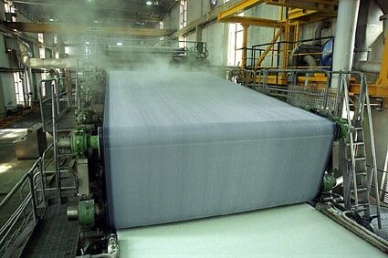 Máquina de fábrica de papel processando um grande rolo de papel, com vapor subindo.
