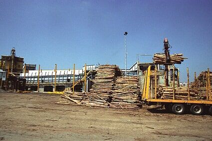 Depósito de madeira com pilhas de tábuas, um caminhão sendo carregado e prédios industriais ao fundo, sob um céu azul.