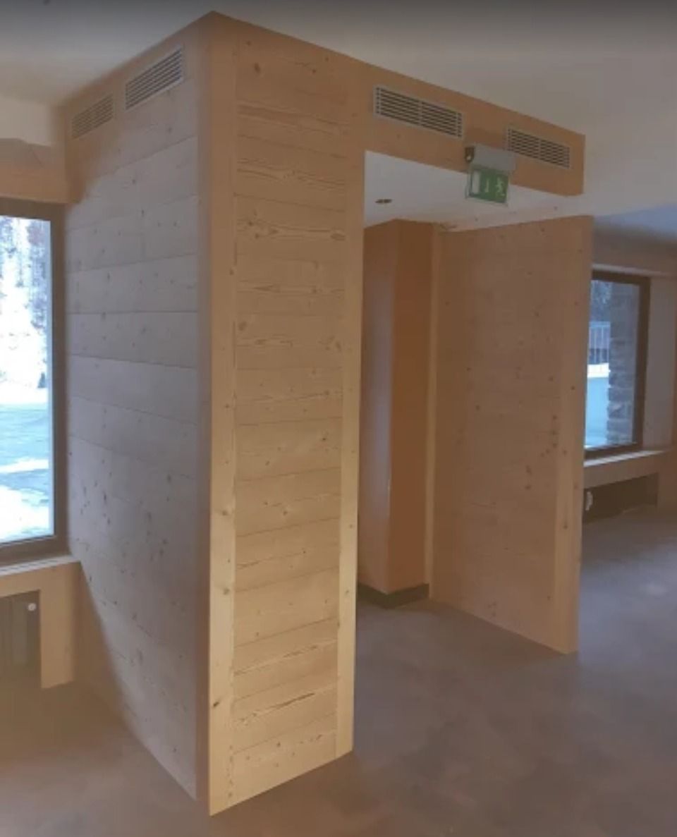 Entrée en bois avec murs à plusieurs niveaux, grilles d'aération et panneau de sortie. Fenêtres et lumière naturelle en arrière-plan.