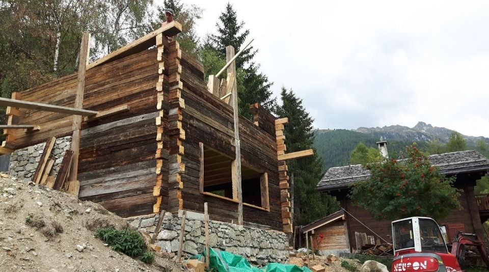 Cabane en bois en construction dans un cadre montagneux, bois brun, fondations en pierre, ciel couvert.